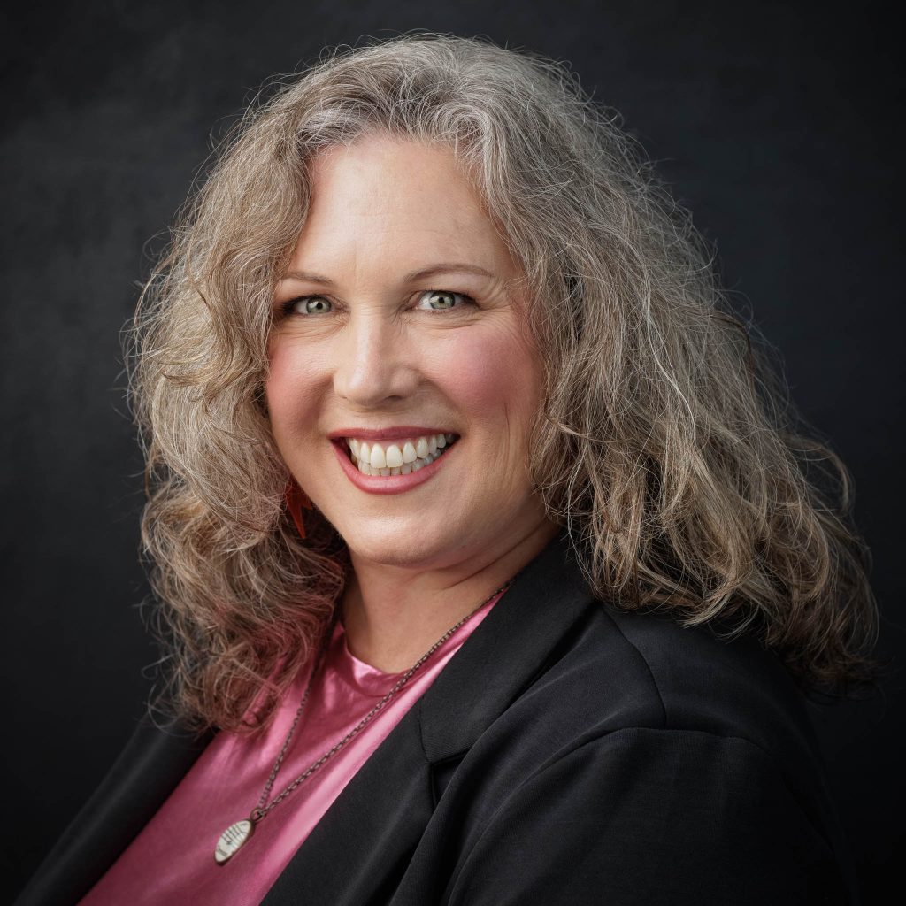 A white woman with brown gray mid-length hair smiles at the camera wearing a black blazer and pink shirt and a long pendant necklace. She is against a dark background.
